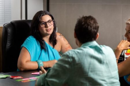 Importações de remédios à base de cannabis crescem mais de 100% a cada ano 4 Imagem de uma mulher branca com cabelo escuro na altura do ombro, sentada numa mesa e conversando com alguém à sua frente. Psicóloga entrevistada para a matéria sobre Importações de remédios à base de cannabis.